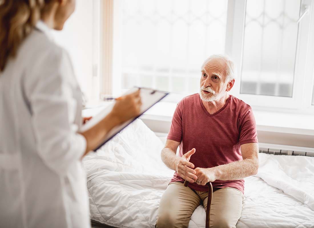 Hospital Indemnity Plans - Senior Male Patient with Cane Talking about His Health with a Female Physician Who is Taking Notes in the Patient’s Hospital Room